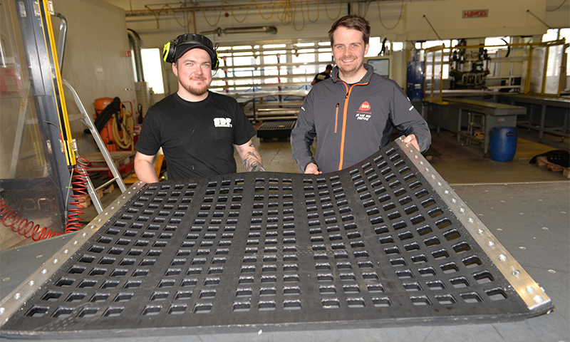 To arbeidere i lyst industriverksted holder et stort svart panel med rutenettmønster.