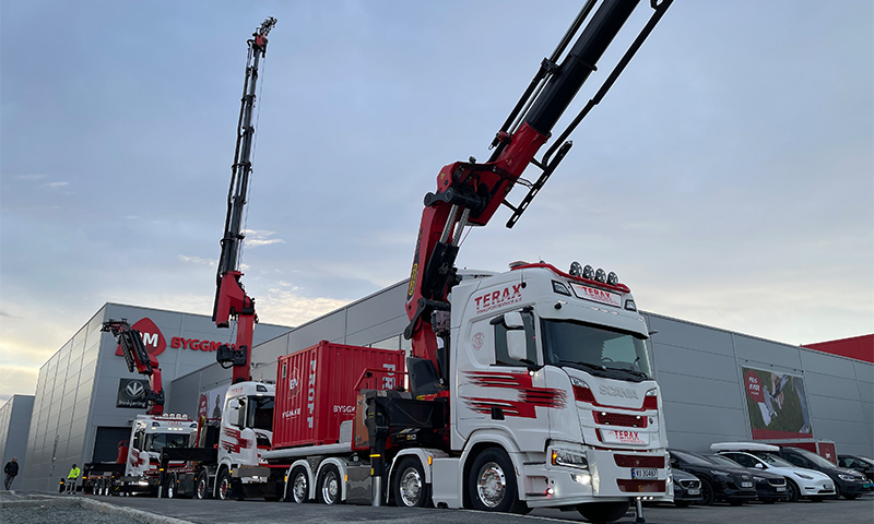 To hvite Scania-lastebiler med røde kraner løfter røde containere utenfor grå industribygning.
