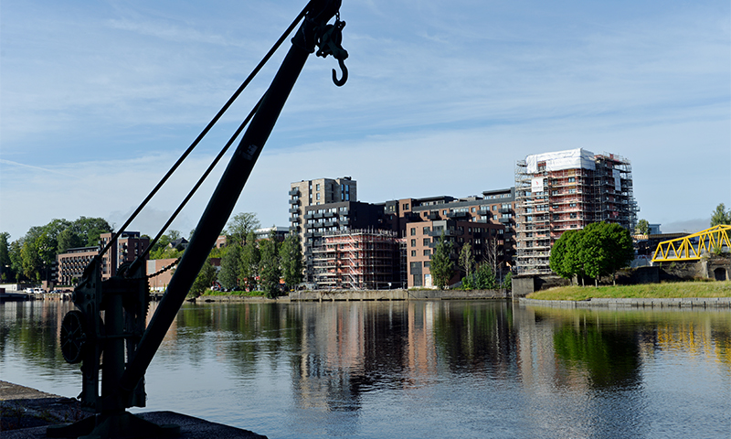 Svart kran på elvebredd, stille vann speiler brune boligbygg under oppføring, gul bro og blå himmel
