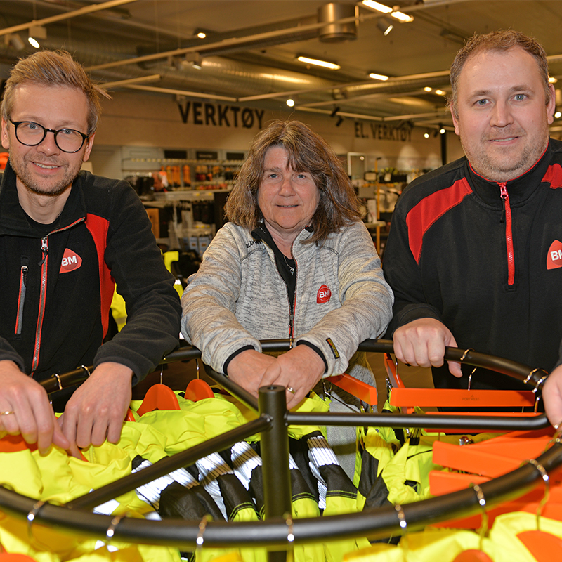 Tre ansatte bak rundt stativ med gule refleksvester i verktøyavdeling.