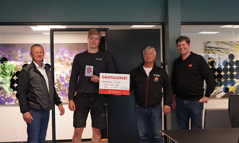 Fire menn foran blå vegger og lilla bilde, en holder skilt med “Gratulerer!” i rødt-hvitt.