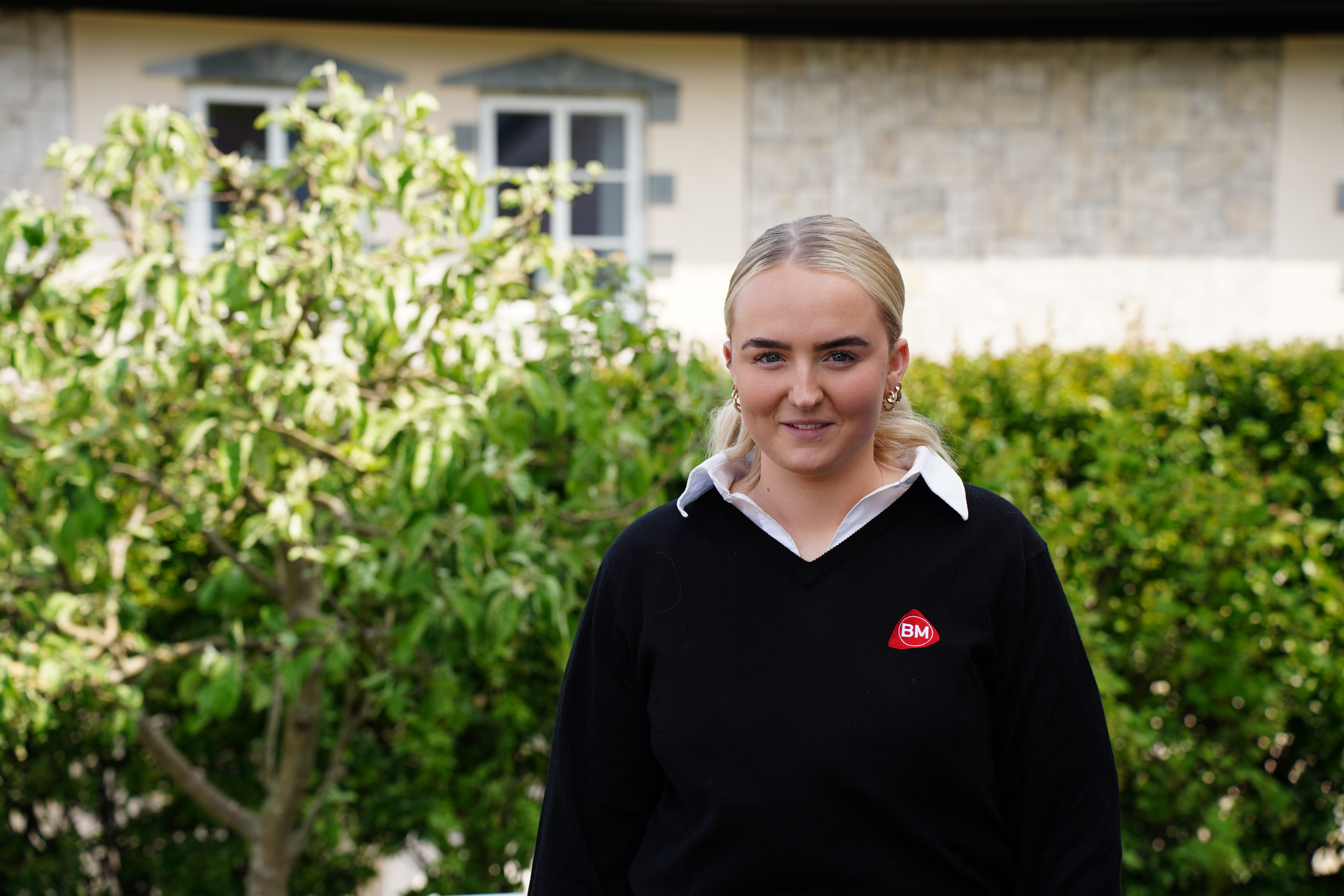 Blond person i svart genser med hvit skjortekrage og rødt BM-emblem foran grønn hekk og bygning.