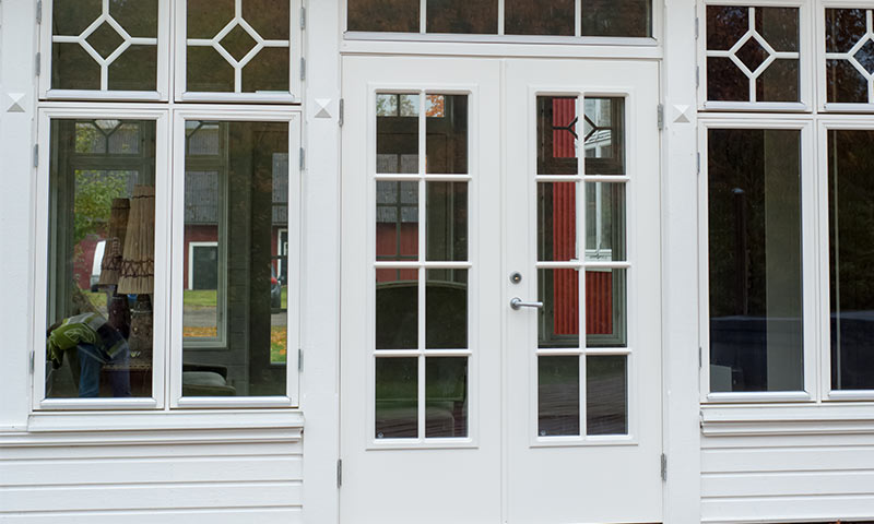 Hvite dobbeltdører med sprosserte ruter, flankert av vinduer, speiler rødt hus og grønn plen