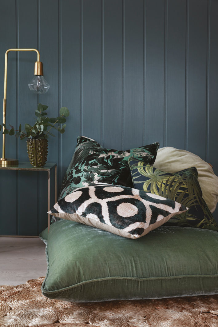 Stablet grønne og mønstrede puter foran mørk blå panelvegg, gull lampe og grønn plante ved siden.