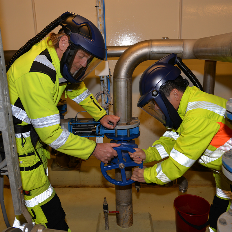 To teknikere i gule verneklær og blå hjelmer vrir et blått ventilhjul i teknisk rom.