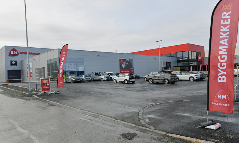 Grått Byggmakker-varehus med røde flagg og parkerte biler under grå himmel.