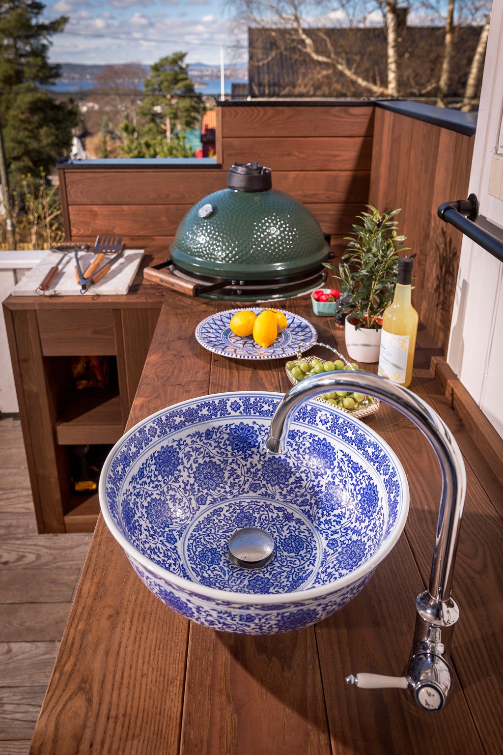 Utendørs benk med blå-hvitt porselensvask, kromkran, grønn grill, sitroner, druer og flaske.