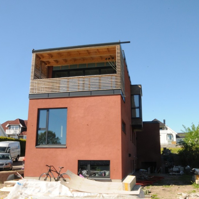 Ny rød funkisbolig med øverste treverkbalkong, store vinduer, sykkel og byggeplass.