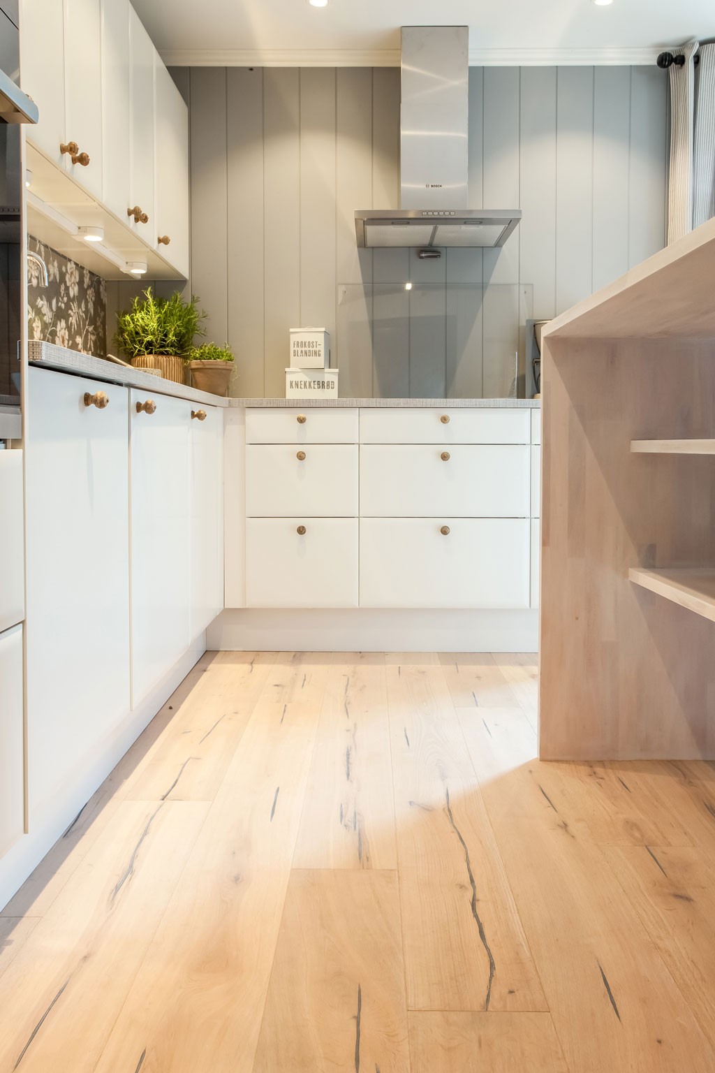 Lyst moderne kjøkken med hvite skap, grå panelvegg, stålhette, planter og lyse tregulv.