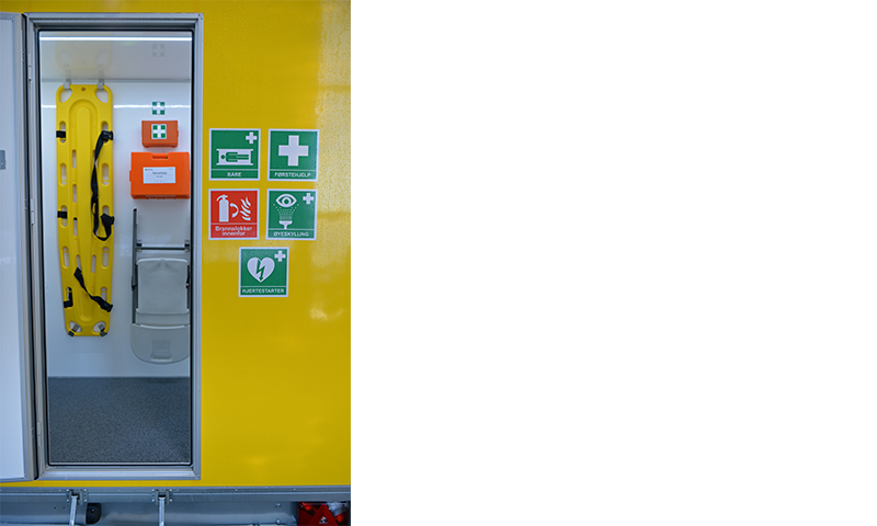 Åpent gult nødhjelpskjøretøy med gul båre, oransje førstehjelpskoffert og grønne sikkerhetsskilt
