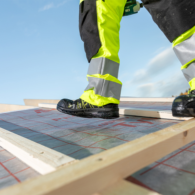 Nærbilde av byggearbeider i neon gule refleksbukser og svarte vernesko på takramme med blå himmel
