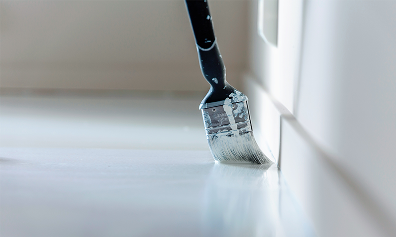 Pensel med svart håndtak maler hvit gulvlist langs vegg
