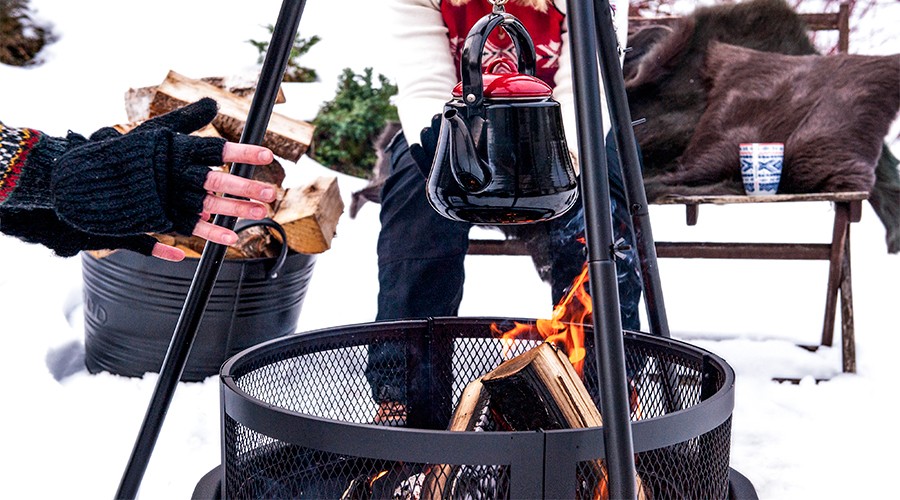 Svart kjele med rødt lokk over bål i snø, votthånd varmes, vedstabel og kopp i bakgrunnen.