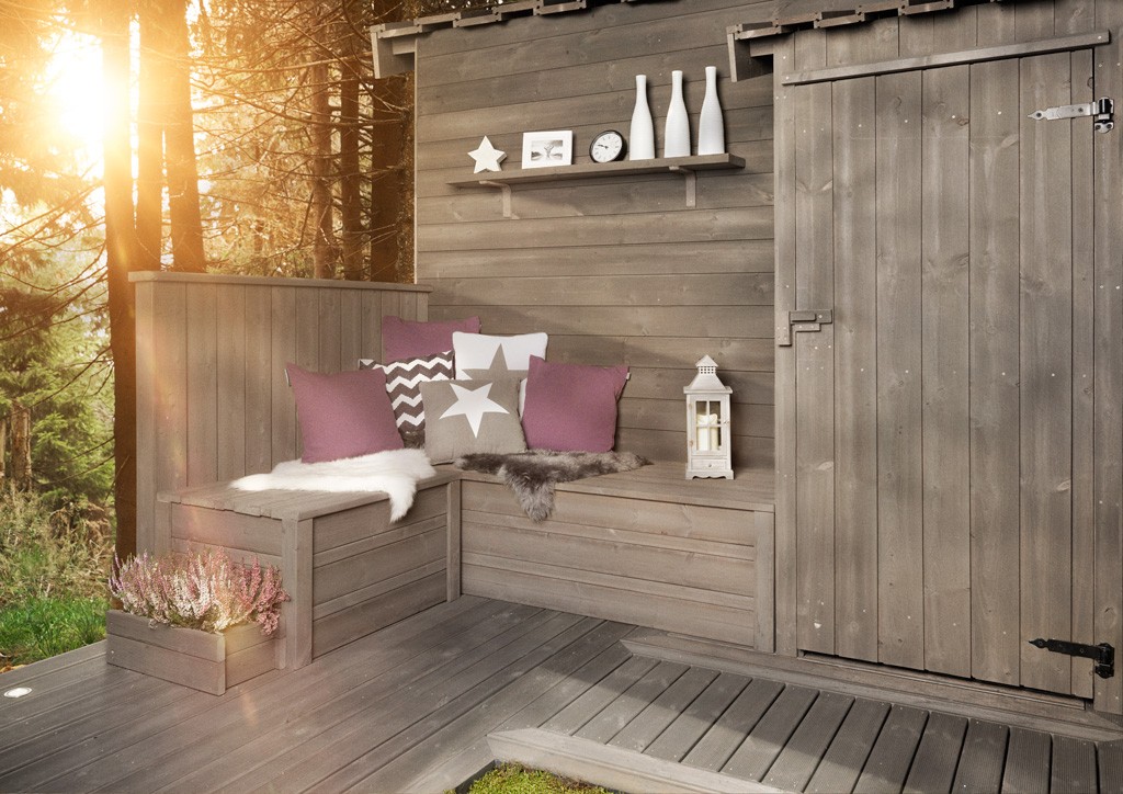 Grå hjørnebenk i tre med rosa og grå puter, lammeskinn, lykt på terrasse mot solfylt skog.