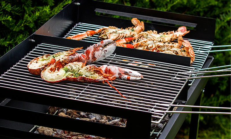 Halvdelte røde hummer med urtefyll på svart grill utendørs mot grønn hekk.