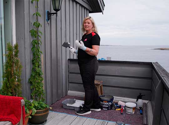 Person maler grå hyttefasade på veranda ved sjøen, med malingsspann og grønne planter rundt