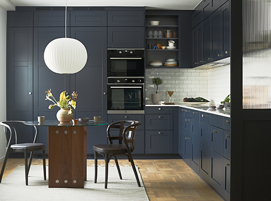 Moderne kjøkken med mørkeblå skap, hvit flisvegg, glassbord, hvit pendellampe og blomstervase