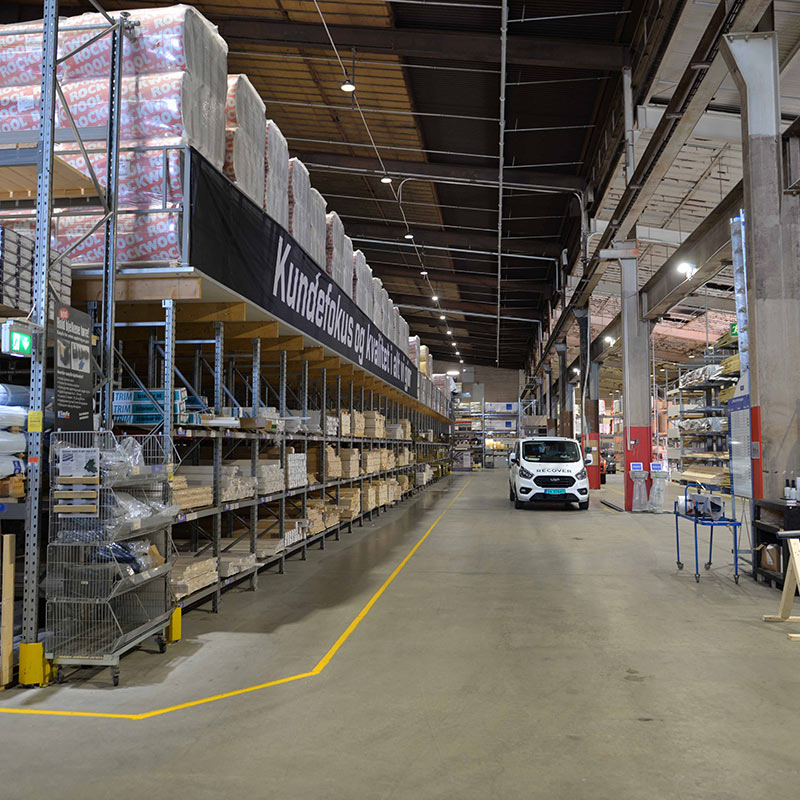Stor lagerhall med høye hyller av byggevarer, hvit varebil i midtgangen og gule gulvmarkeringer.
