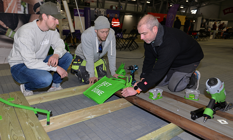 Tre håndverkere på messe monterer gulvplanker med grønne verktøy og handlenett «GET IT DONE»