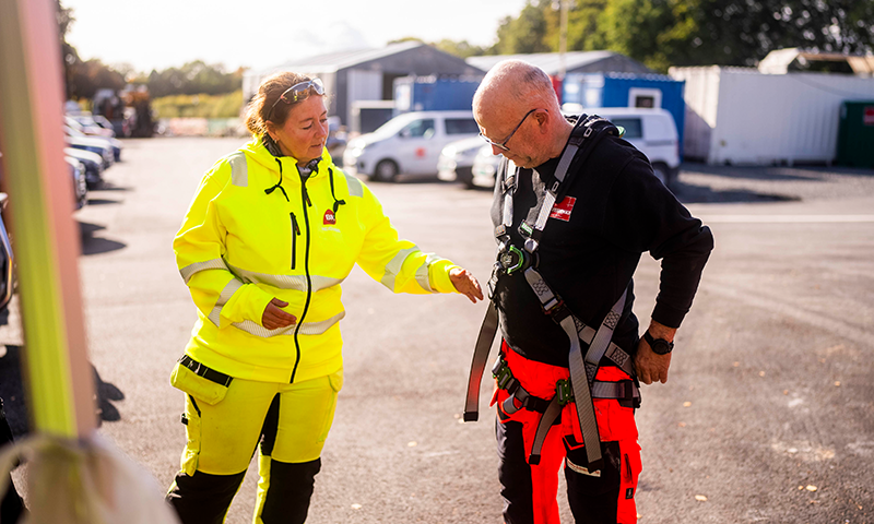 To arbeidere ved byggeplass med containere, gul jakke og svart genser med rødt sikringssele