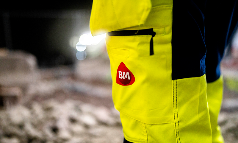 Nærbilde av gul vernebukse med rødt BM-logo, byggplass i nattemørke