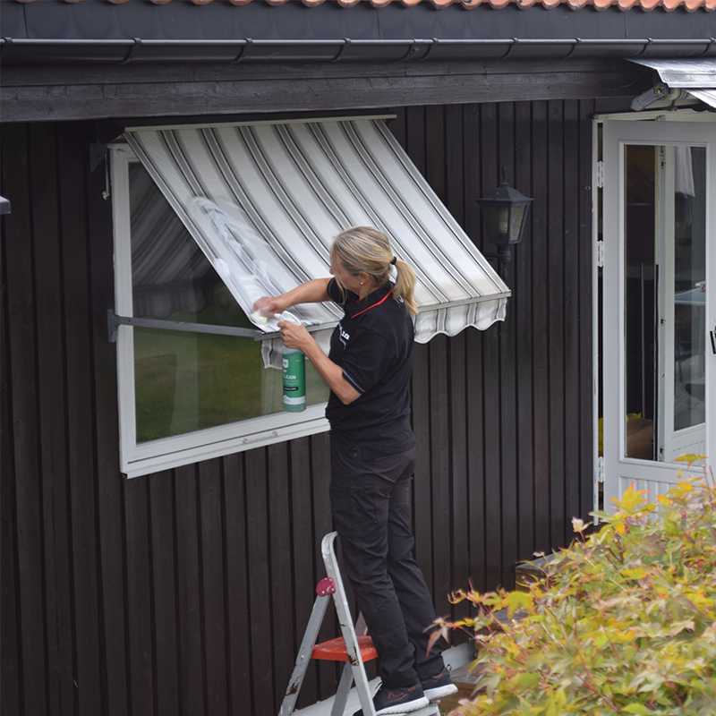 Kvinne i svart uniform står på stige og vasker vindu på mørk trebygning med stripet markise.