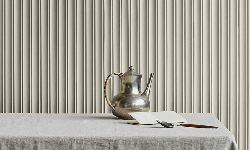 Sølv tekanne med flettet håndtak på grå duk, ved åpen bok og treskje foran beige plissert forheng.