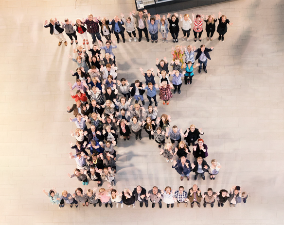 Oversiktsbilde av fargerike mennesker som danner bokstaven K på lyst flisgulv, vinker