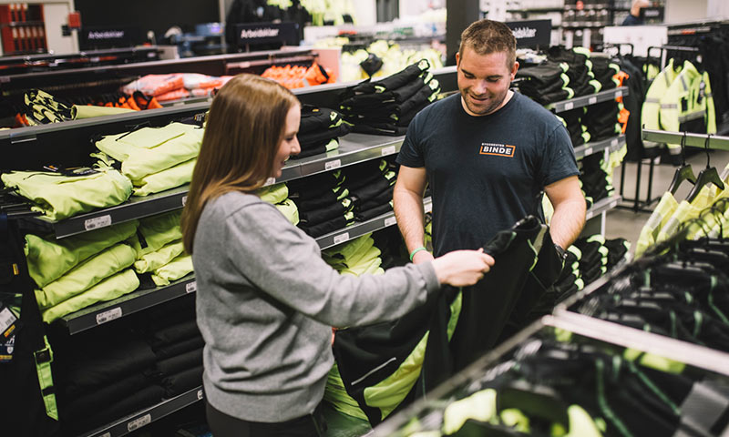 En kunde og ansatt i en arbeidsklærbutikk holder svarte og gule refleksplagg foran hyller.