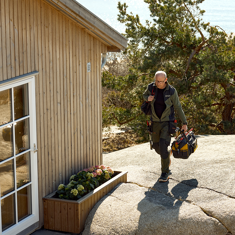 En person i arbeidsklær går mot en trehytte med verktøykasse i hånden. Blomsterkasse ved inngangen og trær i bakgrunnen.