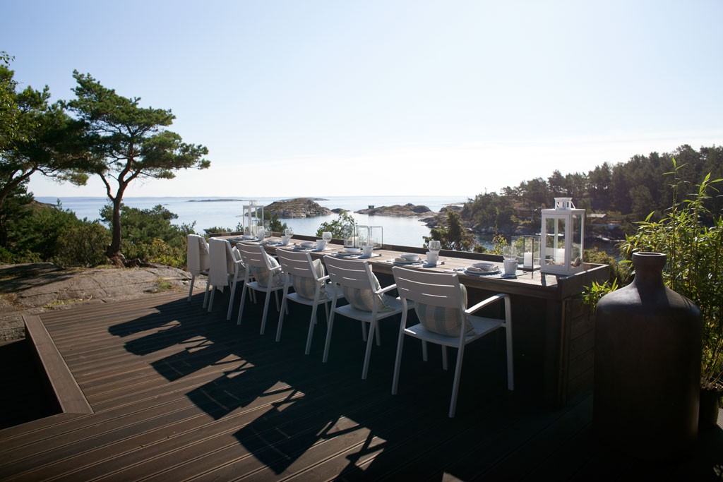 Kystterrasse med langt trebord, hvite stoler og lanterner, utsikt mot svaberg og hav