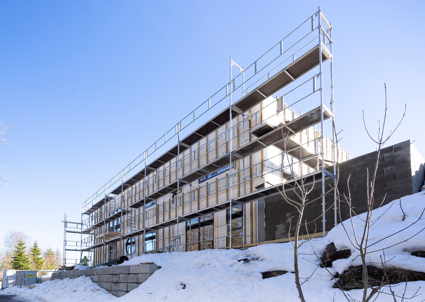 Fleretasjes bygg under oppføring med stillas, trepanel, snødekt bakke og klar blå himmel.