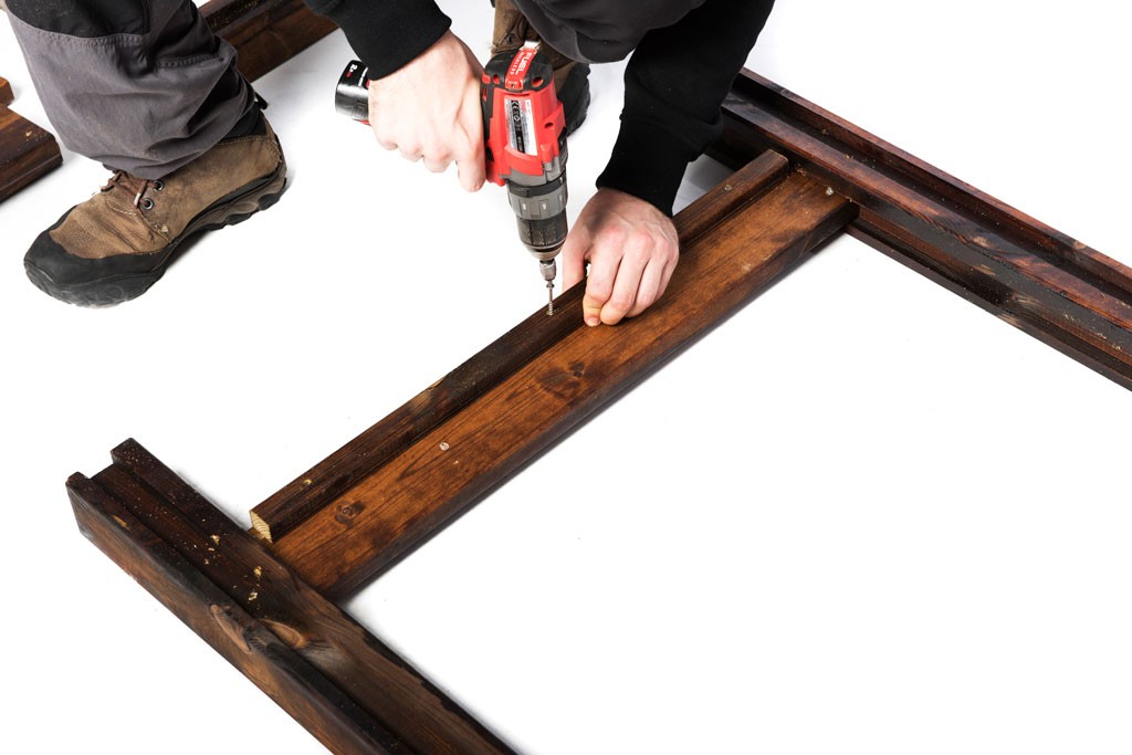 Nærbilde av hender med rød drill som skrur mørk brunt treverk på hvit bakgrunn