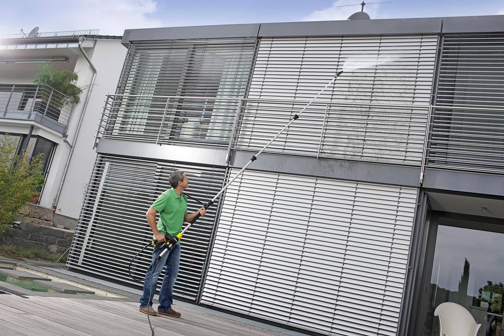 Mann i grønn skjorte rengjør grå fasadepersienner med høytrykkspyler på moderne hus.