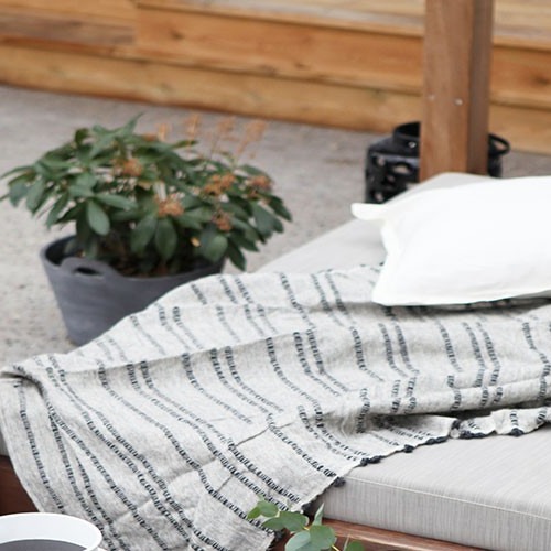 Uteområde med lysgrå hagemadrass, gråstripet pledd, hvit pute og grønn plante.