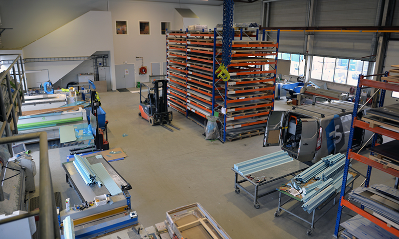 Lyst industriverksted med blå-oransje stålreoler, gaffeltruck og metallplater på vogner