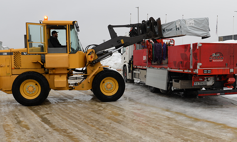 Gul hjullaster løfter hvitt pakkegods mot rød lastebil på isete bakke.
