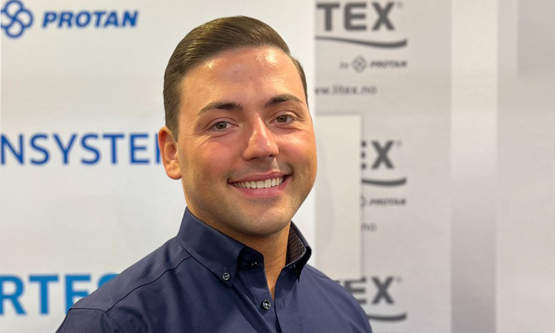 Mann i mørk blå skjorte foran hvit messevegg med blå Protan- og Tex-logoer