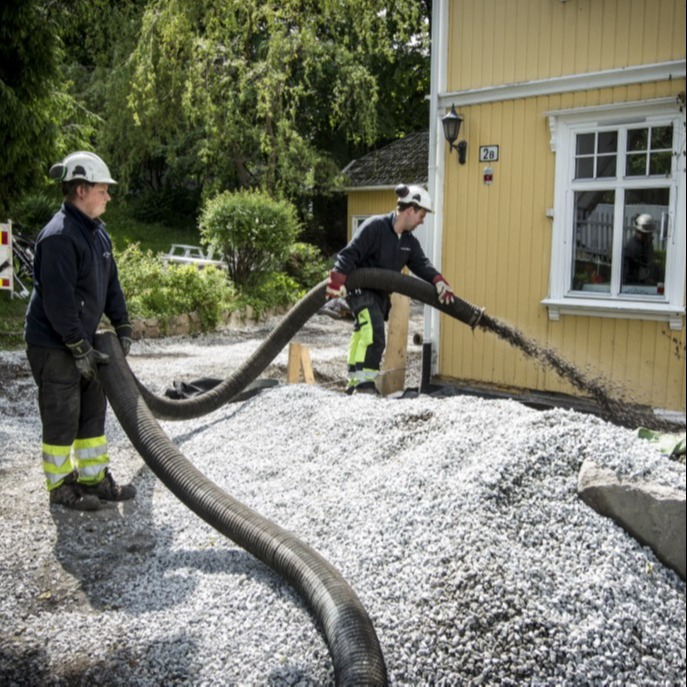 To arbeidere med hjelmer pumper grå pukk med svart slange ved gul trehusvegg.