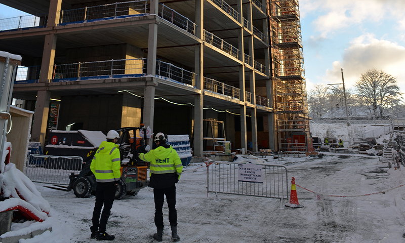 Snødekket byggeplass med fleretasjes betongskjelett i gyllent sollys, to arbeidere i gule jakker