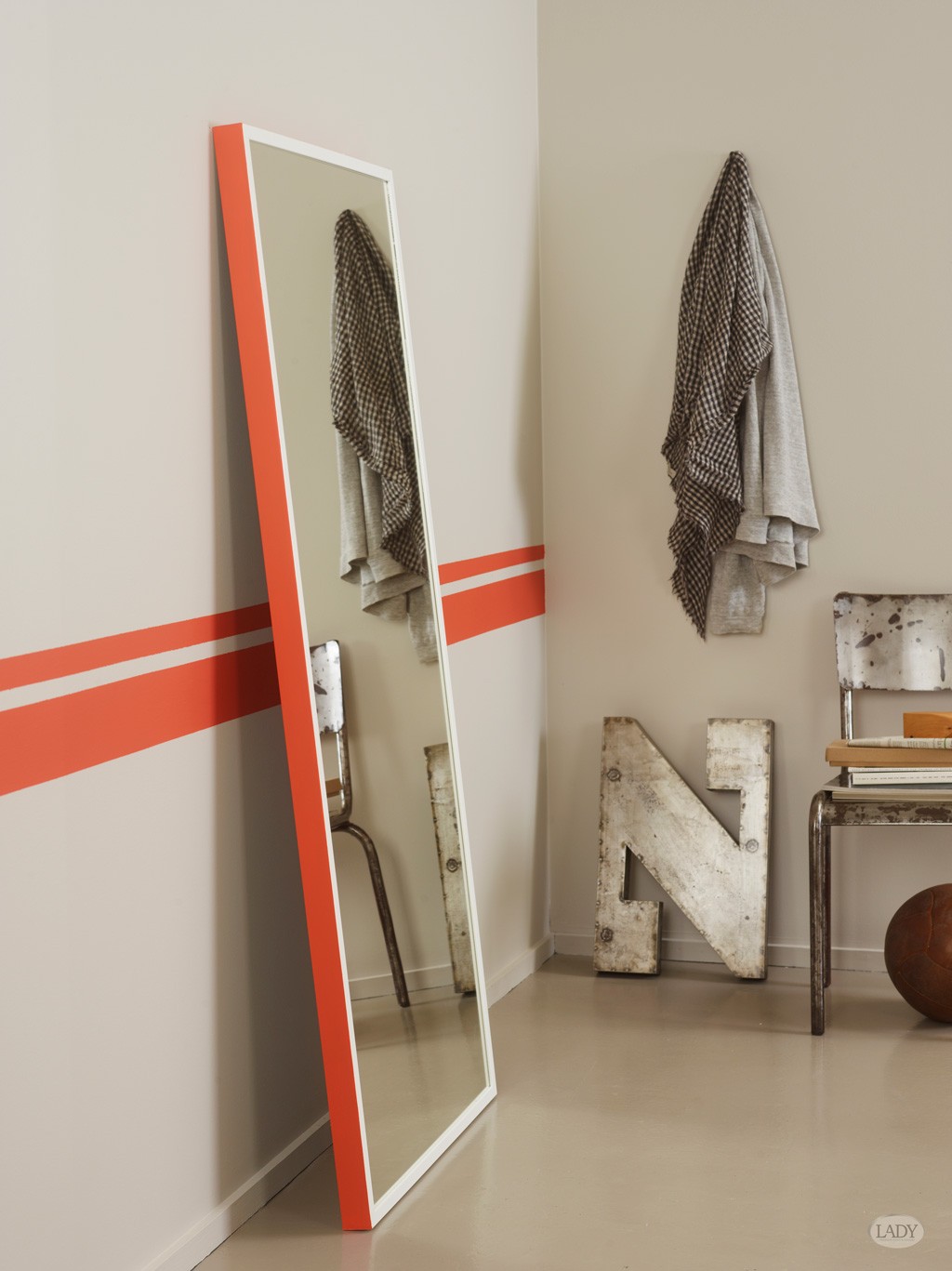 Høyt speil med rød ramme i beige hjørne med røde striper, hengende grå tekstiler og metallbokstav N