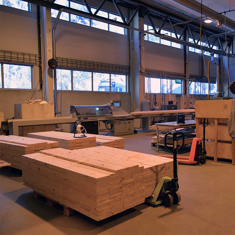 Industrihall med stabler lyse treplanker, maskiner, håndtrucker og høye vinduer mot snølandskap