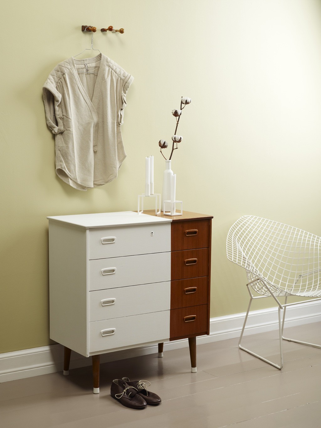 Beige skjorte på krok over tofarget kommode med hvite lys, vase, brune sko og hvit trådstol