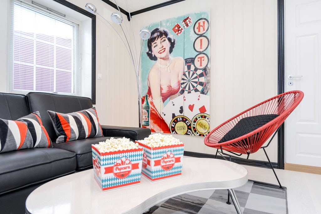 Oppholdsrom med svart sofa, røde/grå puter, hvitt popcornbord, rød lenestol og retroveggkunst.