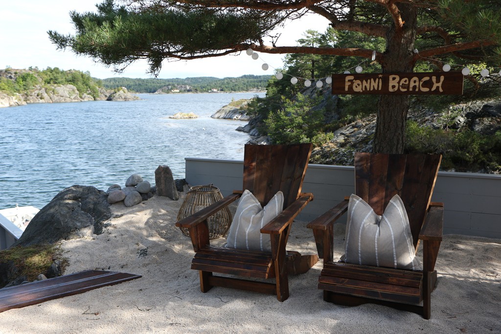 To mørke trestoler med grå stripete puter på sand under furu ved fjord, skilt ’Fanni Beach’