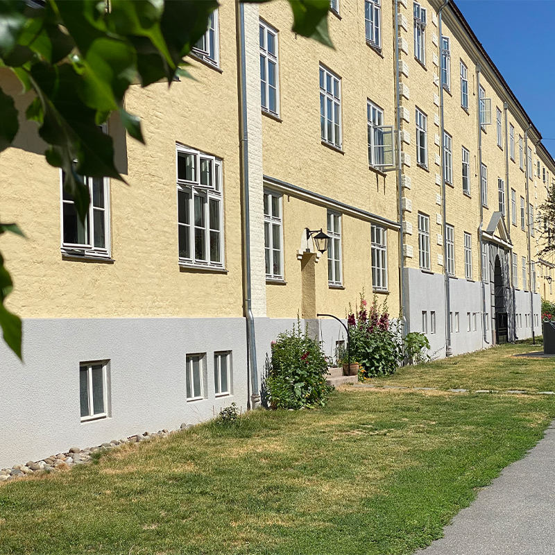 Gult murbygg med hvit sokkel, mange hvite vinduer, grønn plen, blomsterbed og blå himmel.