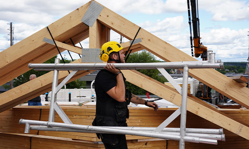 Arbeider med gul hjelm bærer stillasramme foran trekonstruksjon og blå himmel.