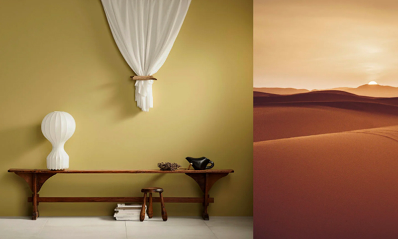 Lang trebenk mot olivengrønn vegg med hvit lampe, hengende stoff, tekanne og ørkenbilde.