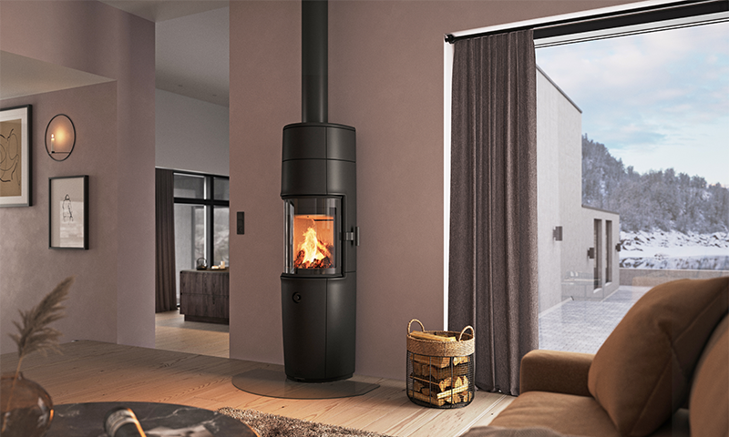 Koselig stue med svart sylinderovn, flammer, vedkurv og stort vindu mot snølandskap.