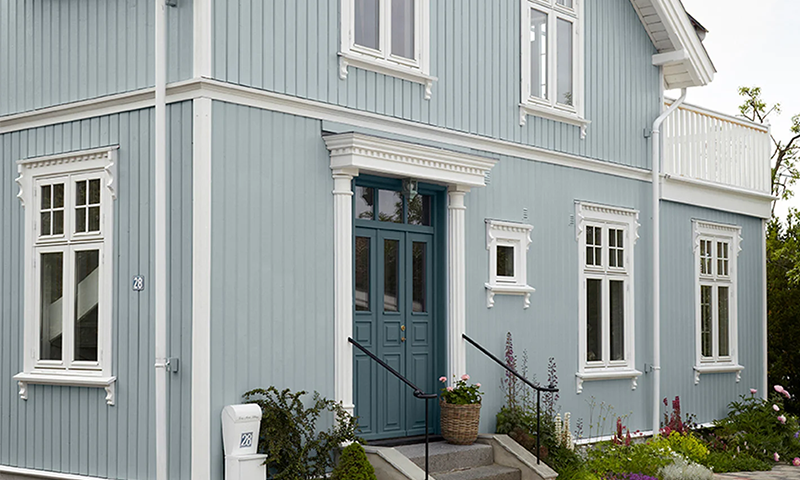 Lyseblå toetasjes trehus med hvite karmer, turkis dobbeltdør og blomster ved trapp